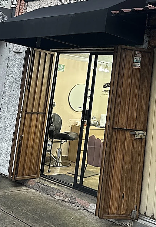 Entrada del local con las puertas de madera abiertas, toldo negro y, detrás del vidrio, el salón con espejos redondos y sillones rosas.